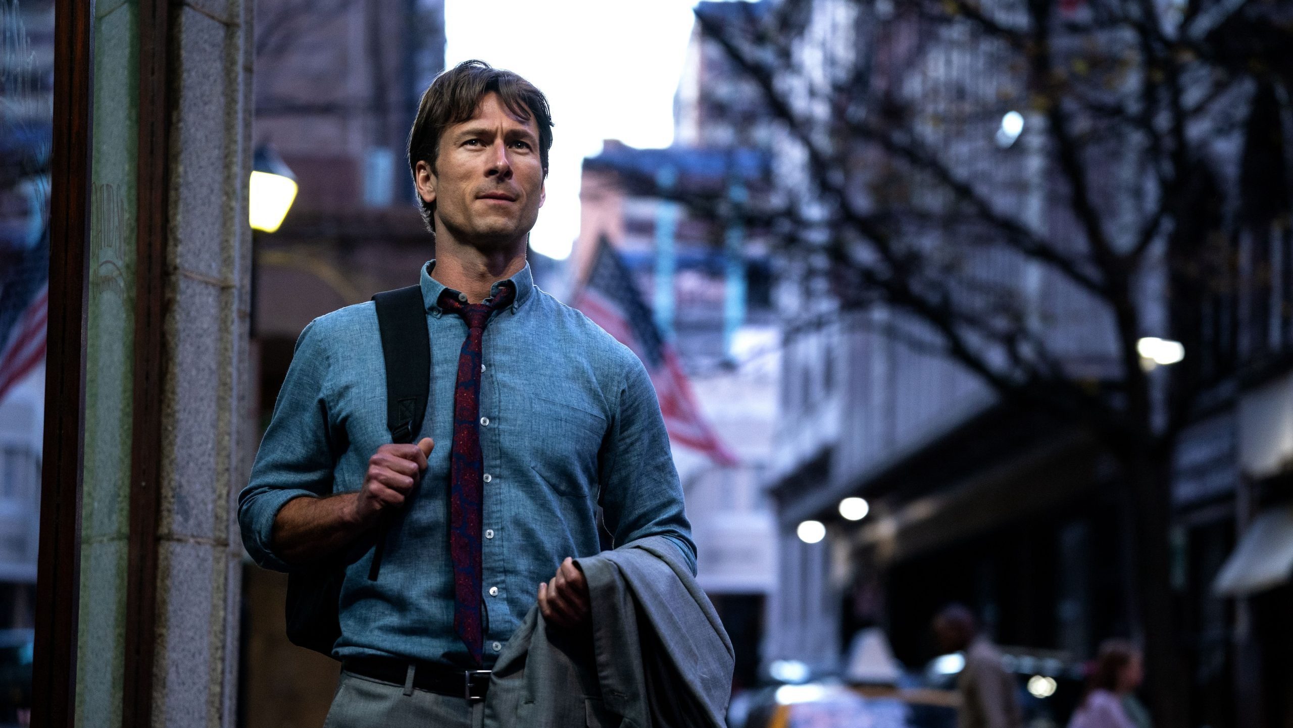 An image from the film How to Make a Killing. It features a man (Glen Powell) wearing a light blue shirt and a patterned tie walks down a city street in the evening, holding a grey suit jacket over his arm and wearing a backpack.
