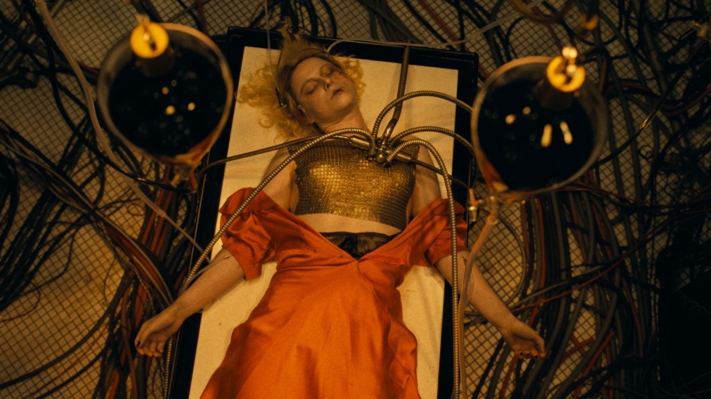 An image from the film The Bride! It features a woman (Jessie Buckley) lying on a futuristic operating table, surrounded by wires and glowing instruments, wearing a metallic top and flowing red skirt.