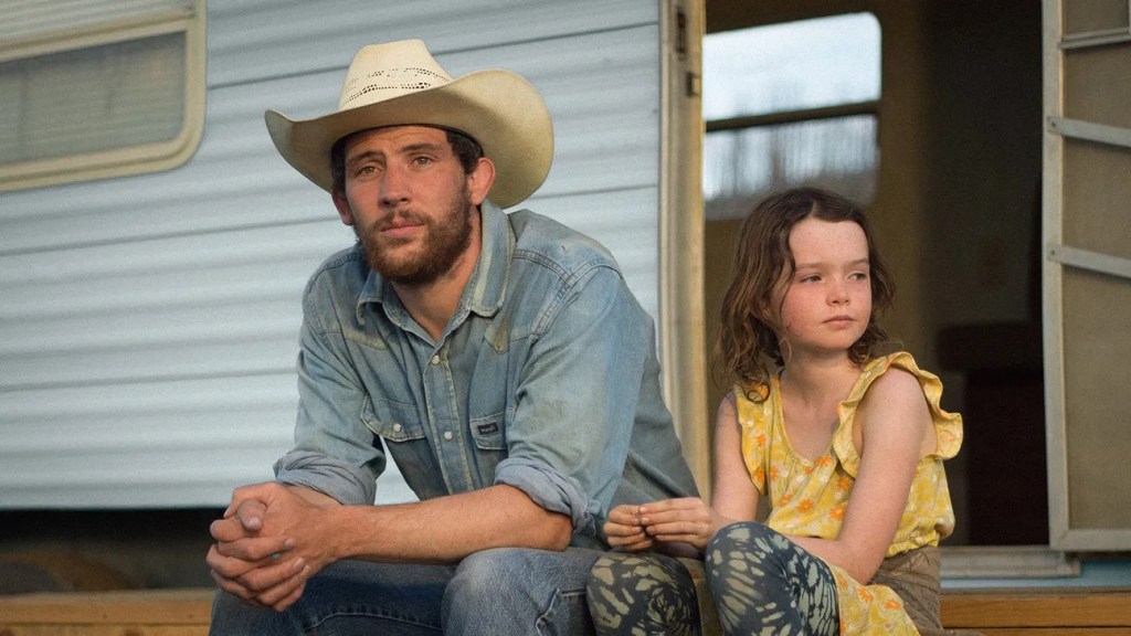 An image from the film Rebuilding. A man (Josh O'Connor) in a cowboy hat and a denim shirt sits beside a young girl (Lily LaTorre) in a yellow dress, both looking contemplative while perched on the steps of a trailer.